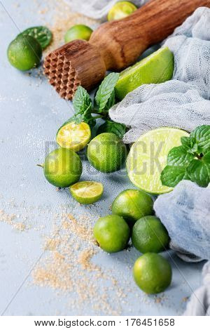 Ingredients for mojito cocktail, whole, sliced lime and mini limes, mint leaves, brown crystal sugar over gray stone texture background with gauze textile and wooden bar muddler. Close up, space