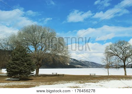 Scenic winter lake landscape with tall bare trees and picnic tables on shoreline of frozen snow covered lake.Bright blue sky and white cloud over mountains in background. Snowy field in foreground.