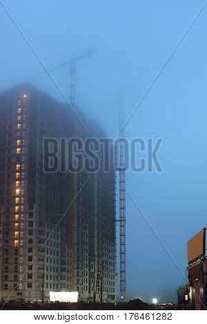 Two cranes on the construction site, unfinished houses, fog covers the upper floors, evening twilight, the lighting of the floors.