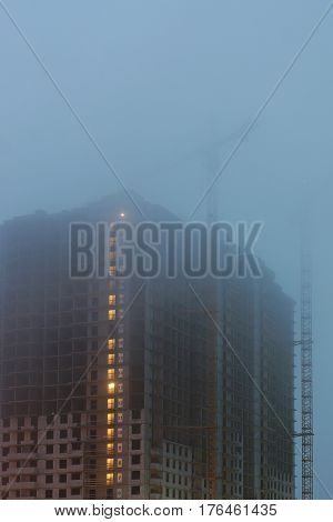 Construction crane on the site, unfinished houses, fog covers the upper floors, evening twilight, the lighting of the floors.