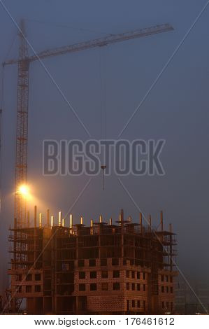 Crane on the construction site, unfinished house, foggy evening twilight, building lighting.