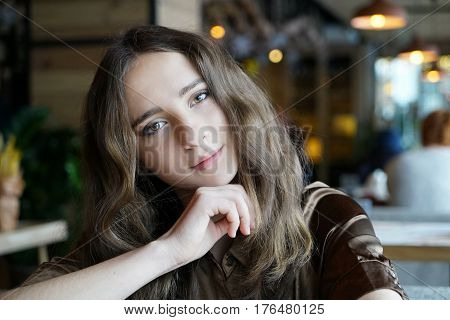 Portrait of young long-haired girl  with bokeh on the backdrop. Curly haired woman sits in coffee shop and smils. Dark-brown eyes, mouth closed, healthy skin.