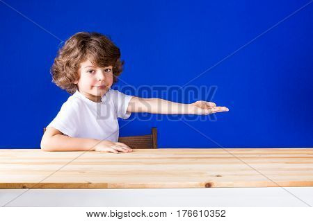 Funny curly cute boy sitting half-turned his hand outstretched and smiling looking at the camera. Close-up. Blue background.