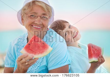 child and grandmother eating watermelon on the beach