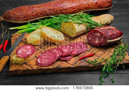 Small antipasto sandwiches from baggetta bread raw smoked sausage on board knife. Ingredients for sandwiches with sausage thyme rosemary fresh onions red hot peppers. Dark black wooden background