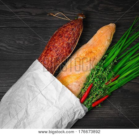 A grocery shopping set in a paper bag at the grocery store for sandwiches. Smoked sausage bread baguette green onion thyme red pepper. Dark brown wooden background.