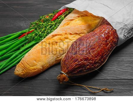 A grocery shopping set in a paper bag at the grocery store for sandwiches. Smoked sausage bread baguette green onion thyme red pepper. Dark brown wooden background.