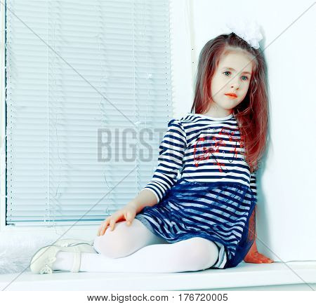 Little girl in blue striped dress and a white bow on her head.She sits at the window with sad eyes.Creative toning of a photograph.
