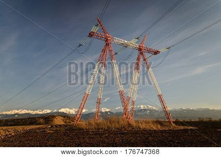 Red and white high voltage tower in the middle of plowed field. Warm late afternoon sunlight with snowy mountains in background and blue sky