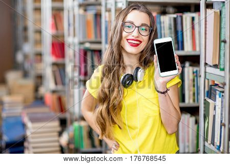 Young smiling student showing smart phone with empty screen standing at the library. Mobile application concept