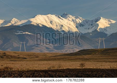 High voltage power towers and lines with snowy peaks of Fagaras Mountains in background a a plowed field in foreground. Horizontal view with warm tones of afternoon sunlight and blue sky.