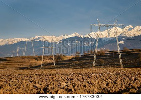 High voltage power towers and lines in the middle of a plowed field. Horizontal view with warm tones of afternoon sunlight snowy mountains in background and blue sky