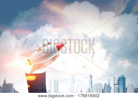 Close up of a hand of a businessman holding a toy rocket and a clowdy cityscape in the background. Toned image. Double exposure