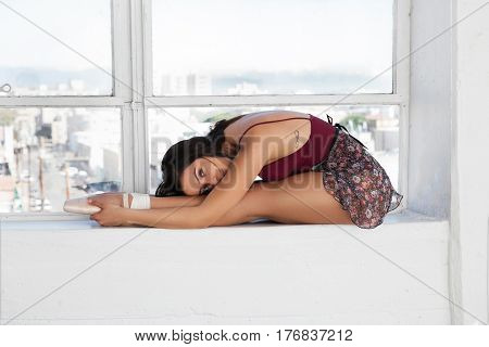 Beautiful young dancer in pointe shoes at white windowsill making ballet leg stretching.