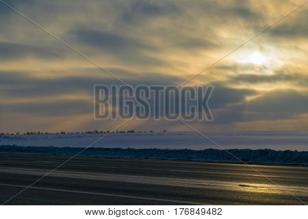 Winter. In the foreground a country paved two lane road. In the background field covered in white snow. The distance the planting of trees. In the sky crimson clouds. Through the clouds and the shining yellow sun.