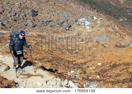 Smiling senior Person in sporty Clothing and with Backpack walking up on steep Mountain Trail from Lodge visible far down
