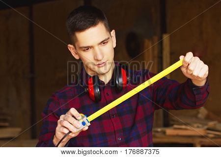 Workman Dressed In The Checkered Shirt Working With Tape-measure At The Sawmill. Timbers On Backgrou