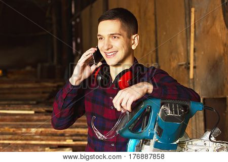 Smiling Workman Dressed In The Checkered Shirt Talking The Phone Near The Circular Saw. Timbers On B
