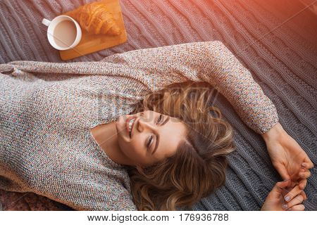 Cheerful woman lying on the bed with the beverage and cornetto.