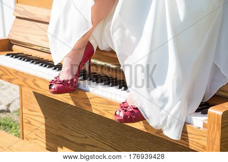 A brides red shoes rest on the keys of a piano that sits outside in the sun