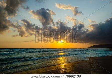 Sunrise over the sea, the rolling calm waves,  horizon is orange, skay orange and blue, clouds are grey and pink.