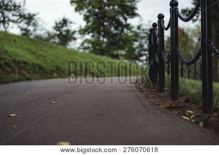 Moody, Dark Photo Of The Road In A Park, Between Woods - Closeup View Of The Chain Fence With Blurre