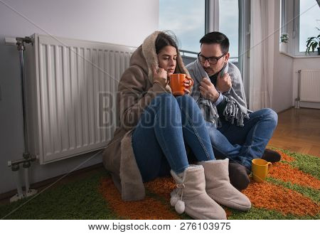 Young Couple In Jacket And Covered With Blanket Sitting On Floor Beside Radiator And Trying To Warm 