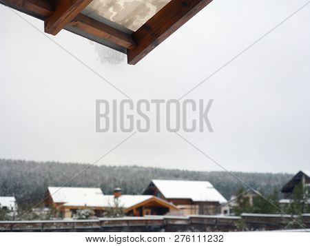 A House Roff With Hanging Snow And Against The Winter Sky, A Village In The Blurred Background