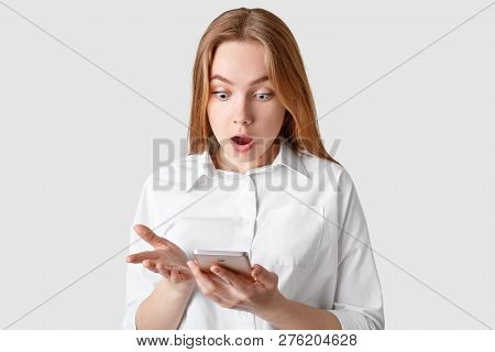 Shocked Terrible Woman Stares At Screen Of Smart Phone, Reads Shocking News, Dressed In Elegant Shir
