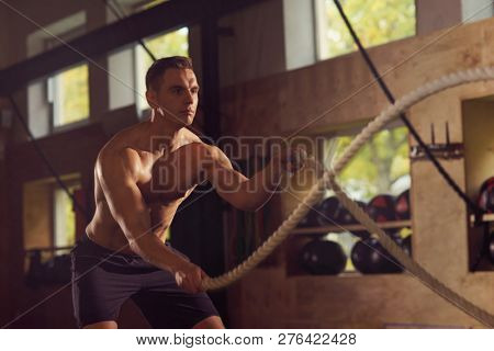 Fit, sporty and athletic sportsman working in a gym. Man training using battle ropes. Sports, athletics and fitness concept.
