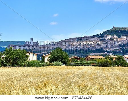 View Of The City Of Assisi, The Slope Of The Mountain Monte Subasio, Summer Clouds, Blue Sky