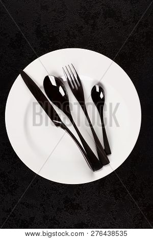 Black Cuttlery On White Plate From Above On Dark Surface. Modern Design Concept.