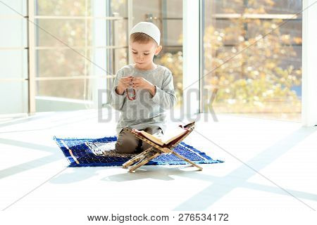 Little Muslim Boy With Misbaha And Koran Praying On Rug Indoors