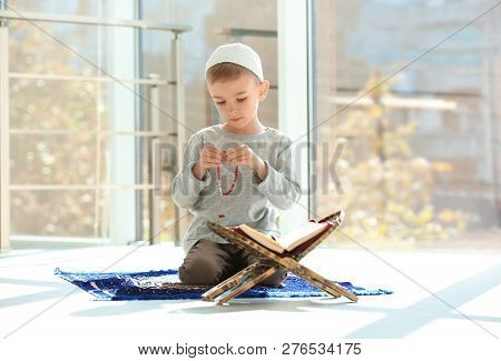 Little Muslim Boy With Misbaha And Koran Praying On Rug Indoors