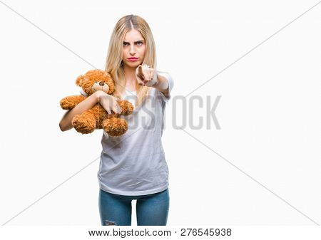 Young beautiful blonde woman holding teddy bear over isolated background pointing with finger to the camera and to you, hand sign, positive and confident gesture from the front