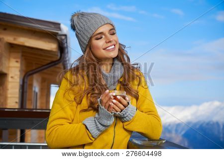 Woman drinking warm tea in the rustick wooden terrace on mountain, alpine view, snow on hills. 