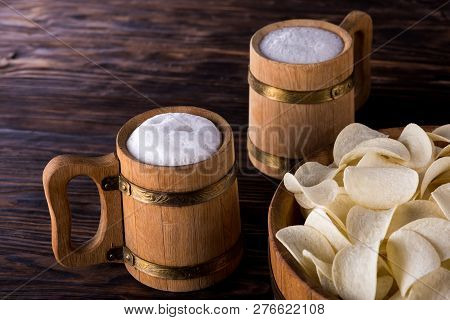 Two Oak Mugs With Beer Inside. They Stand Near The Oak Tray, In Which Are Potato Chips. On Dark Wood