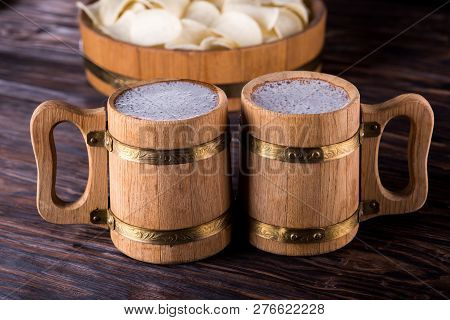 Two Oak Mugs With Beer Inside. They Stand Near The Oak Tray, In Which Are Potato Chips. On Dark Wood