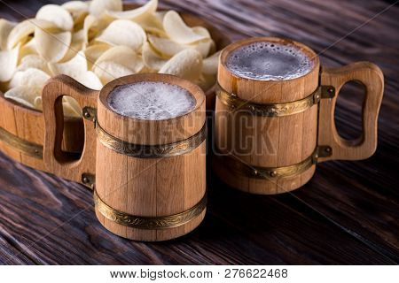 Two Oak Mugs With Beer Inside. They Stand Near The Oak Tray, In Which Are Potato Chips. On Dark Wood