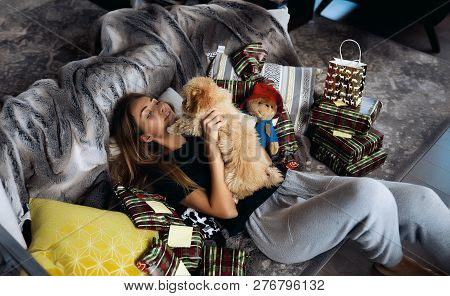 Sweet Couple Of Young Girl And Her Maltipoo Doggy. Lying On Grey Couch With Pillows And Christmas Gi
