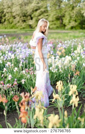 Young European Girl Wearing White Dress Standing Near Irises On Garden.