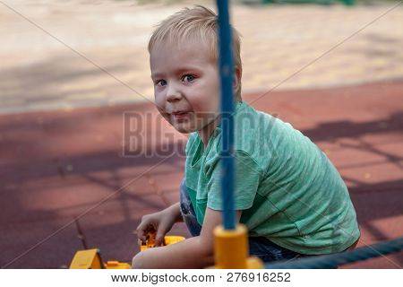 Cute Caucasian Blonde Baby Boy With Blue Eyes Sits On The Cover Of Children Playground With A Toy, Y
