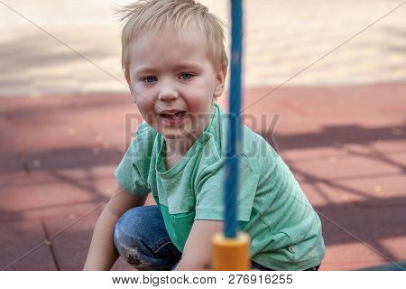 Cute Caucasian Blonde Baby Boy With Blue Eyes Sits On The Cover Of Children Playground. Funny Look, 