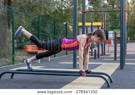 Young Caucasian Woman Workouts On The Park Sports Ground. In Sports Plank Position With One Leg Up, 