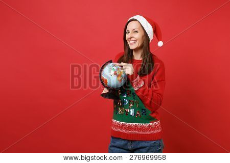 Smiling Young Santa Girl In Knitted Sweater, Christmas Hat Keeping Finger On World Globe In Hands Is
