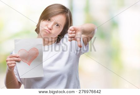 Young adult woman with down syndrome holding red heart card over isolated background with angry face, negative sign showing dislike with thumbs down, rejection concept