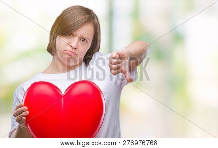 Young adult woman with down syndrome holding red heart over isolated background with angry face, negative sign showing dislike with thumbs down, rejection concept