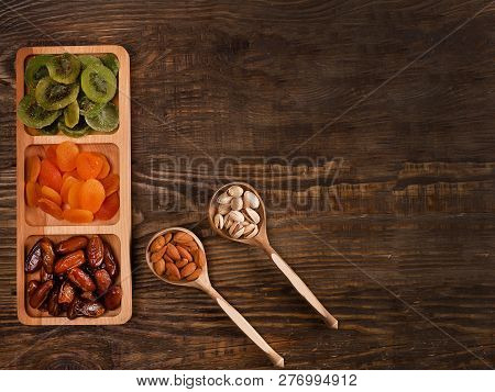 Dates, Dried Apricots And Kiwis In A Compartmental Dish And Nuts In A Wooden Spoon On A Dark Wooden 