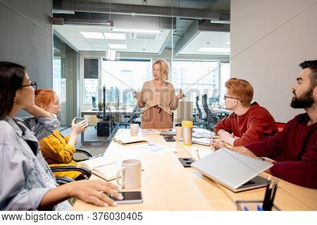 Mature blond woman in casualwear standing by table in front of her young intercultural colleagues during business training in office