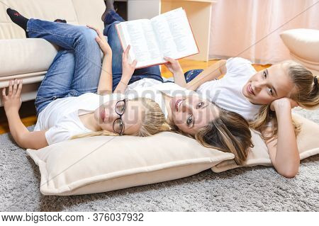 Family Concepts. Portrait Of Positive Mother Reading A Book Together With Her Female Twins Laying On